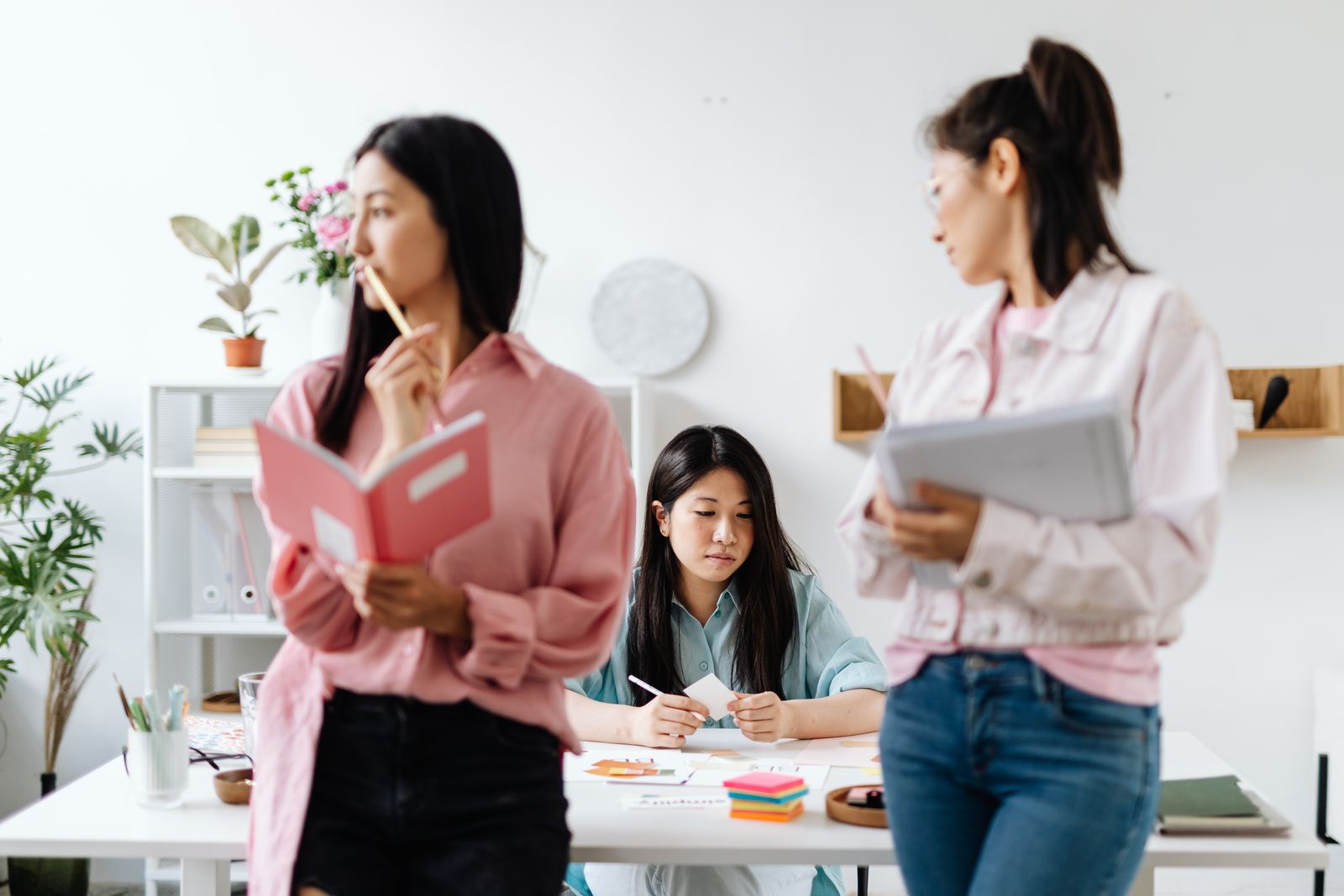 Ba nữ đồng nghiệp trao đổi cạnh bàn làm việc với sổ tay và tài liệu.